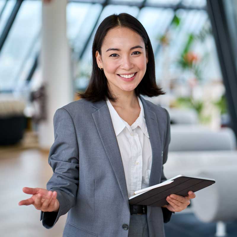 A smiling woman in a gray suit holds a tablet, gesturing warmly in a bright, modern office with large windows and plants in the background.