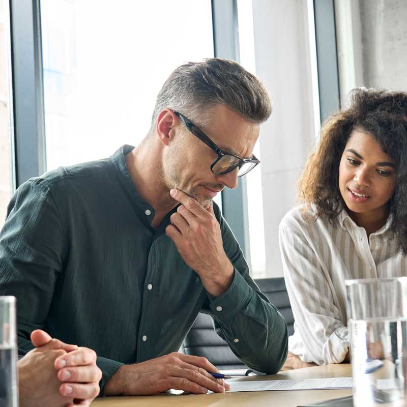 A man and a woman are in a meeting, focused on a document. The man, in glasses and a green shirt, appears thoughtful. The woman, in a striped blouse, listens attentively. Bright office setting.