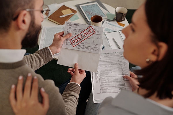 A couple sits worriedly at a table cluttered with bills. The man holds a letter stamped with "Past Due" in red. Papers and coffee mugs are scattered around.