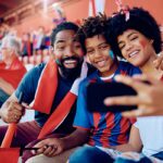 A smiling family of three with face paint takes a selfie at a sports stadium, wearing red and blue team colors while holding a red flag in the stands.