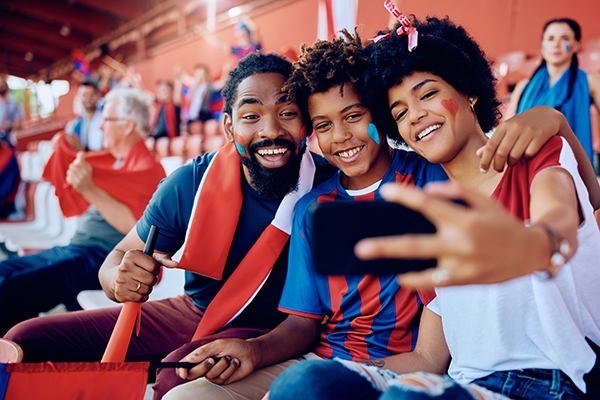 A smiling family of three with face paint takes a selfie at a sports stadium, wearing red and blue team colors while holding a red flag in the stands.