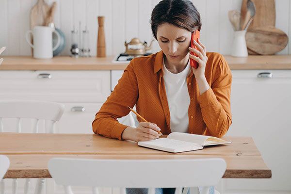 Young woman sits at the kitchen table to make a call while writing notes in a book